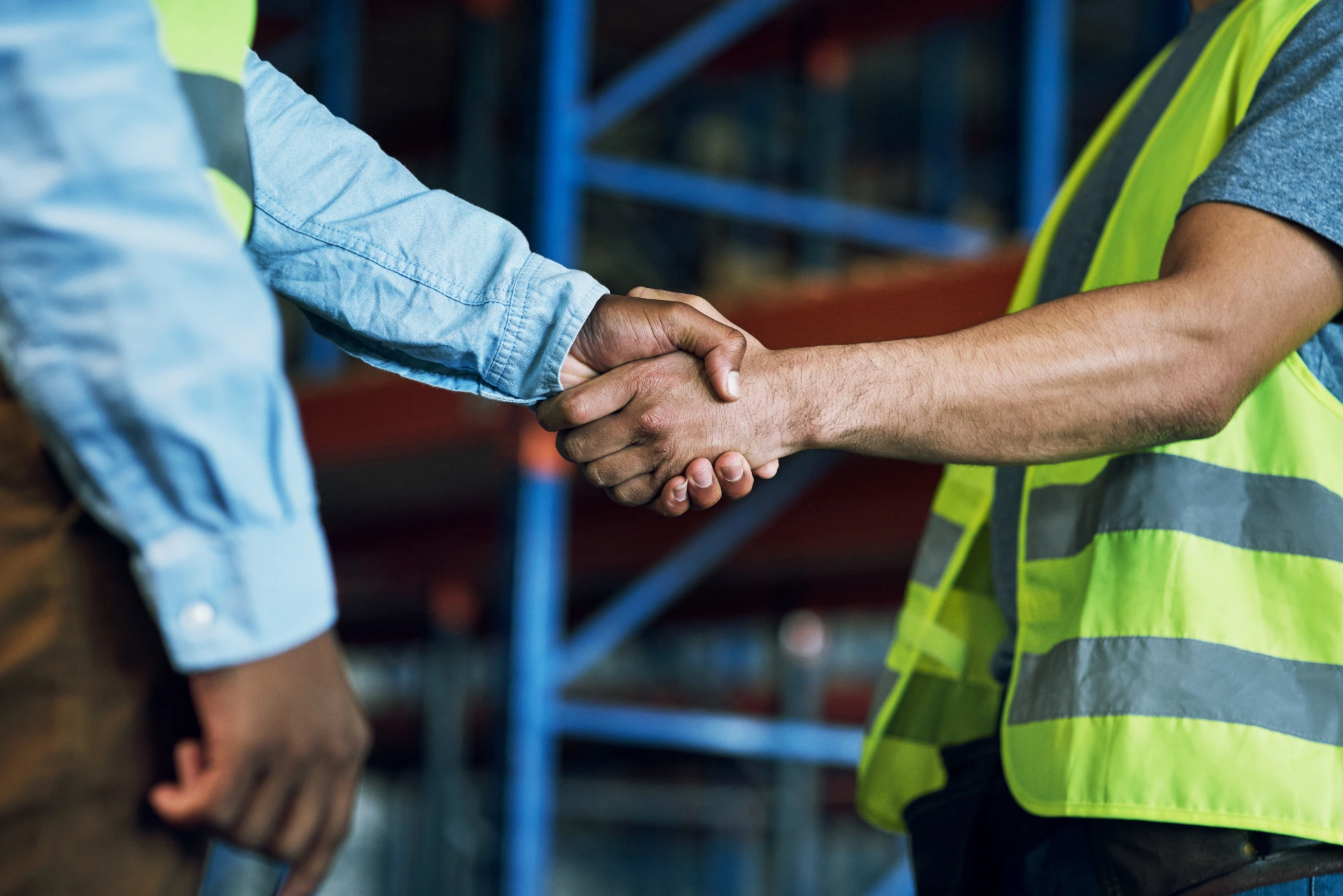 Close‑up of two workers—one in a hi‑vis vest—shaking hands on a factory floor.